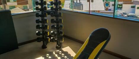 A gym with a bench and dumbbells, large windows with a view of buildings and flags.