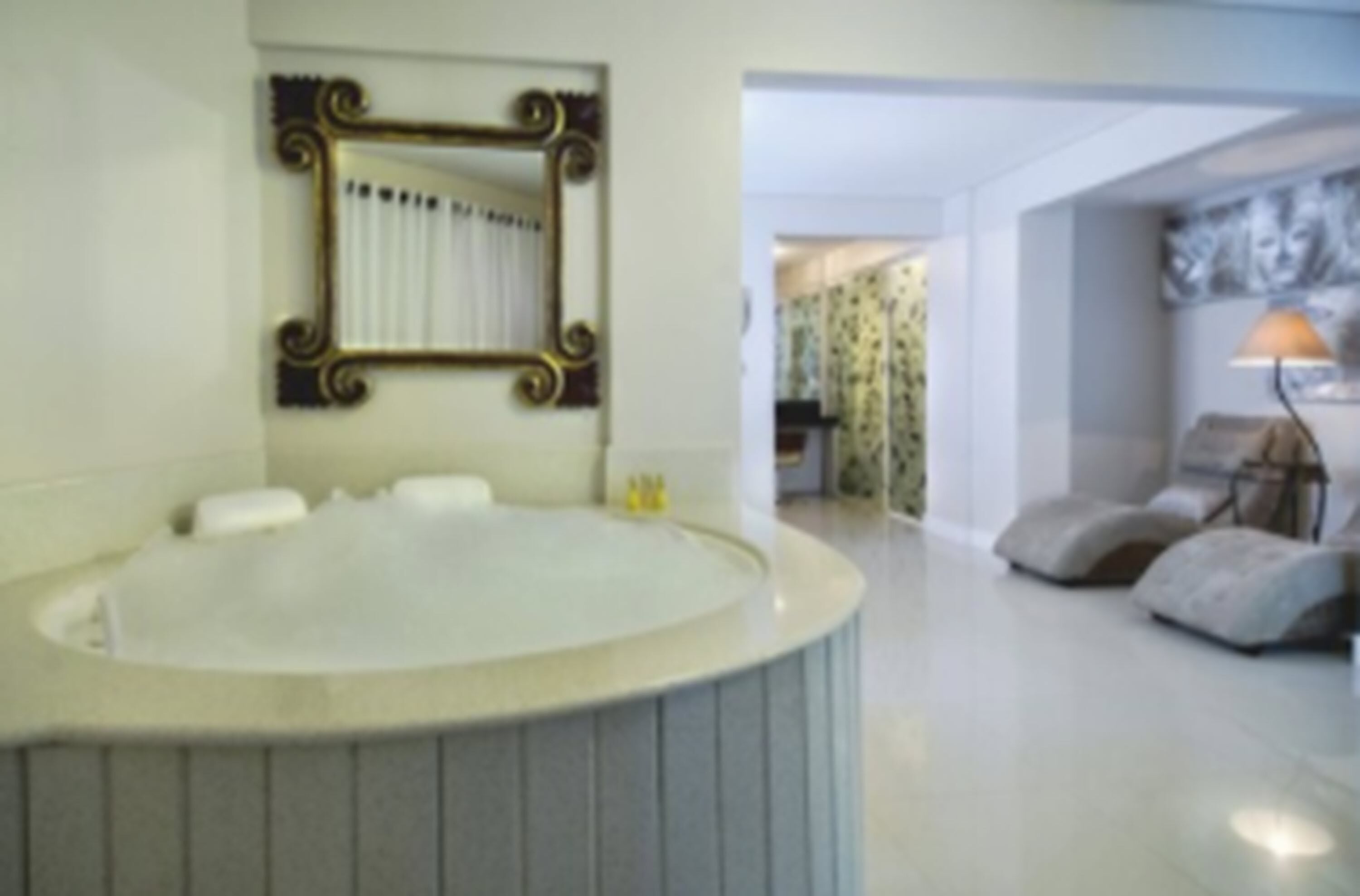 A bathroom with a large bathtub, a mirror with an ornate frame, and a doorway leading to a living area with a sofa and a lamp.
