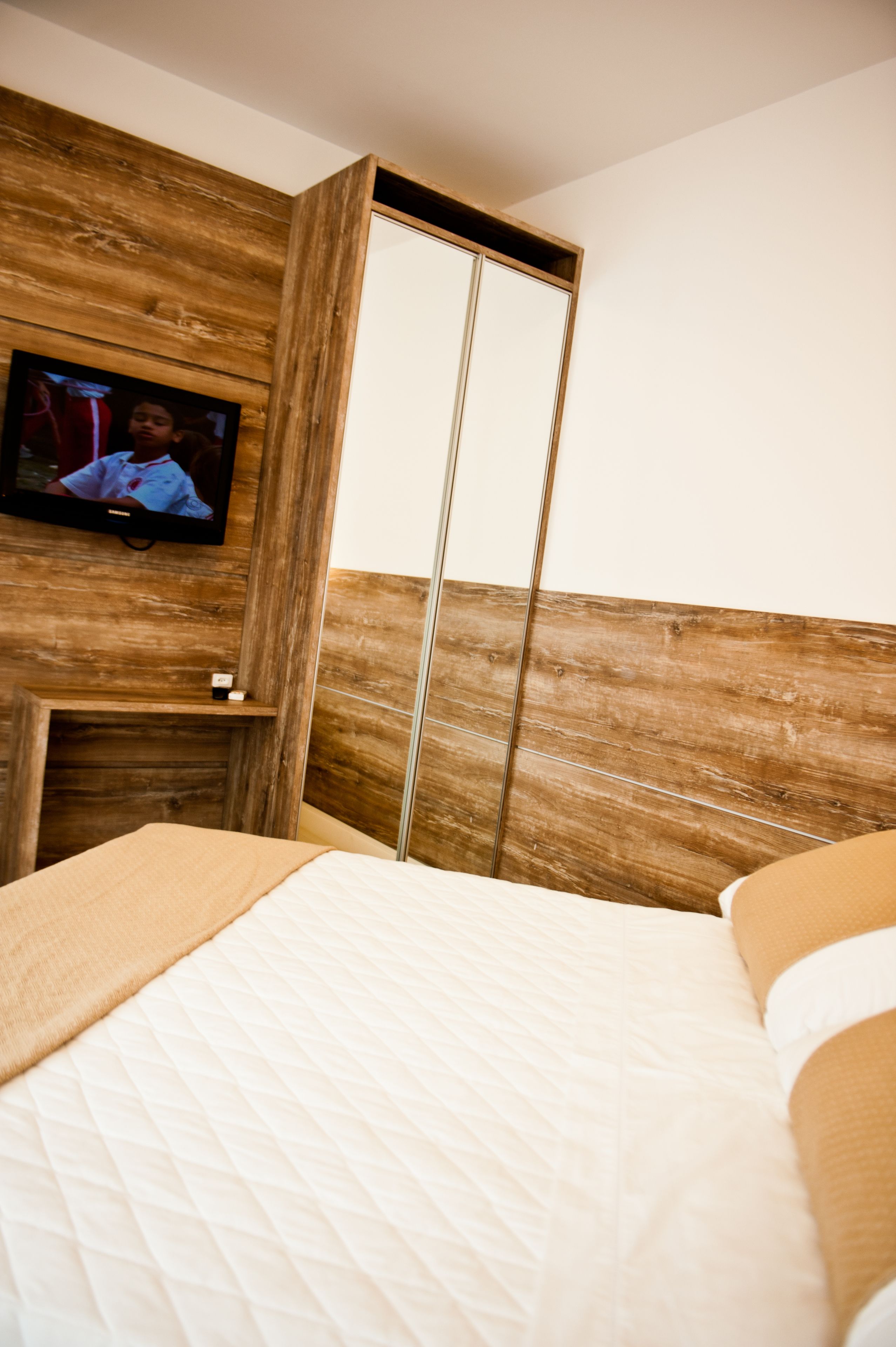 A hotel room with a wooden headboard, a television mounted on the wall, and a bed with a quilted mattress.