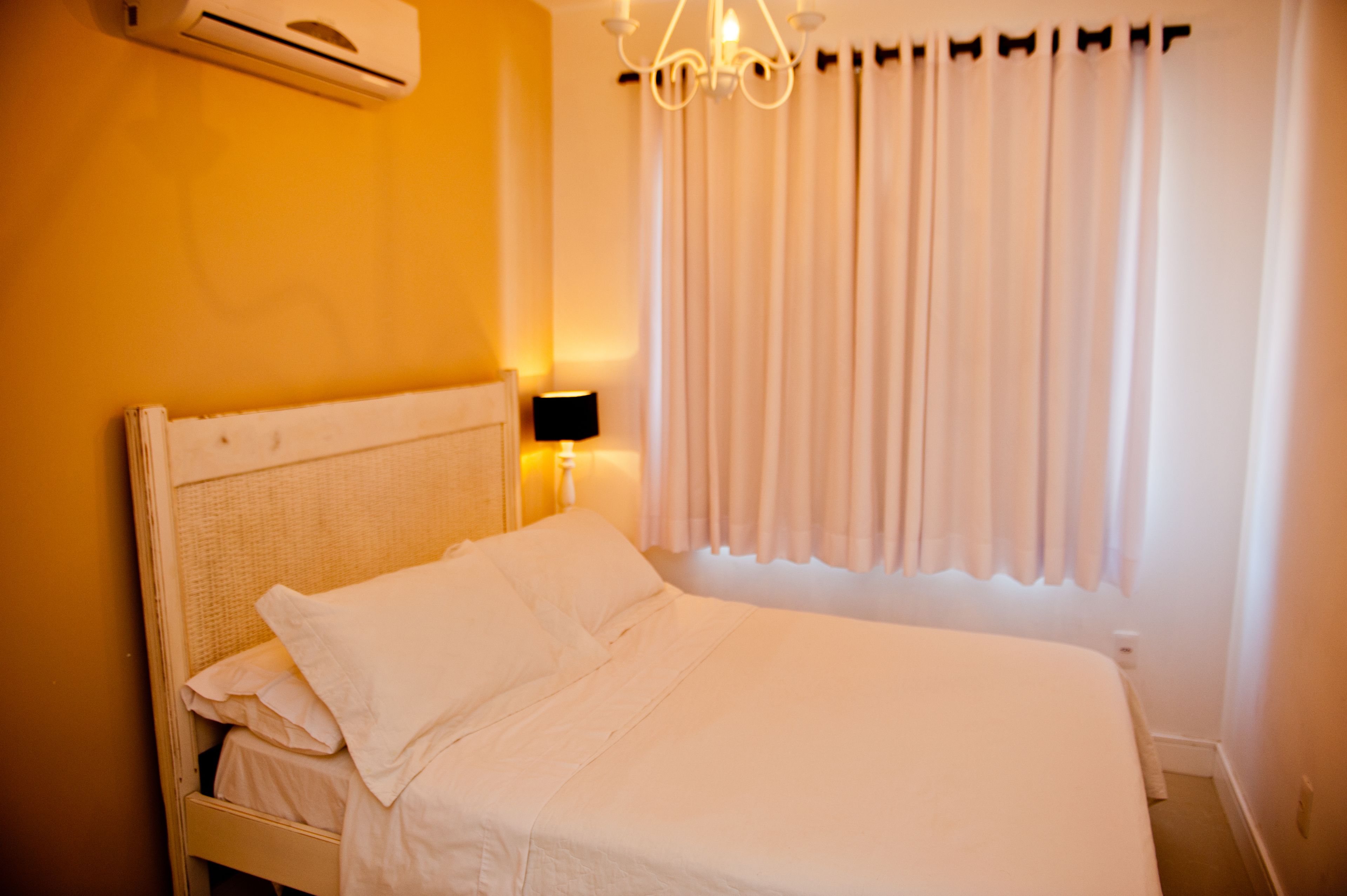Una cama bien hecha con ropa blanca en una habitación con pared amarilla, una ventana con cortinas y un aire acondicionado de pared.