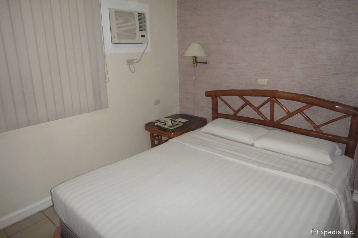 Una habitación de hotel con una cama, una mesita de noche con lámpara, un aire acondicionado instalado en la pared y una ventana con persianas.