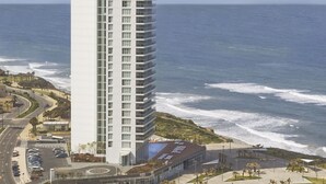 A tall, modern hotel with a white facade and multiple balconies, located near a beach with waves crashing onto the shore.