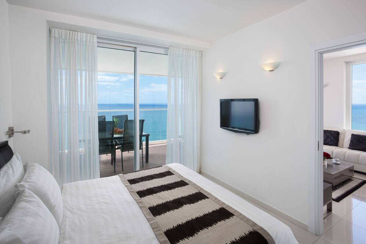 A hotel room with a bed, a TV, a balcony with a table and chairs, and a view of the sea.