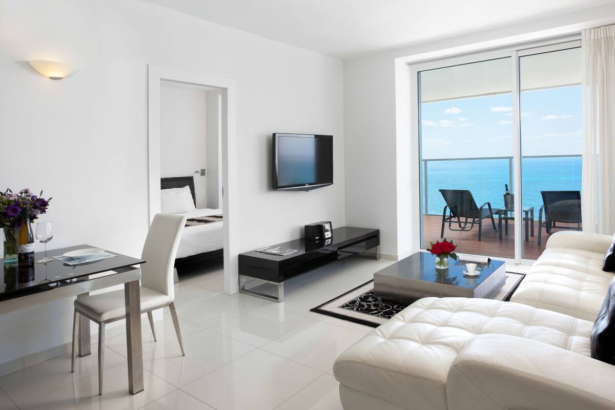 A modern hotel room with a balcony, a flat-screen TV, a sofa, a dining area, and a bed.
