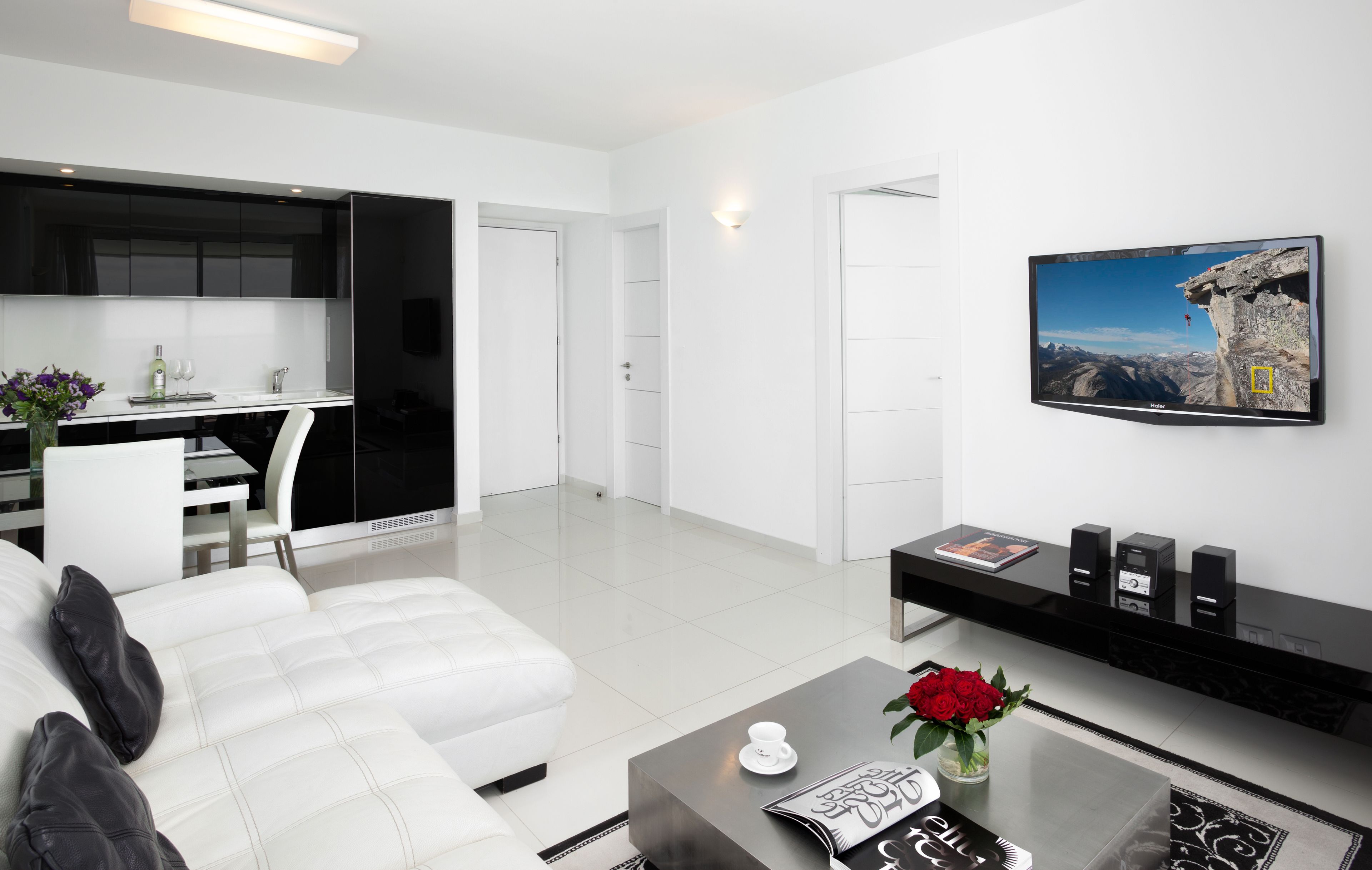 A modern living room with a white sofa, a coffee table with a vase of red roses, a flat-screen TV mounted on the wall, and a kitchen area with black cabinets and a white countertop.