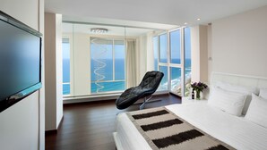 A modern hotel room with a large bed, a black leather chair, a glass wall, and a view of the ocean.