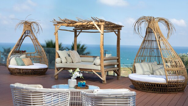A wooden deck with wicker furniture, including a sofa and two hanging chairs, overlooking the ocean.