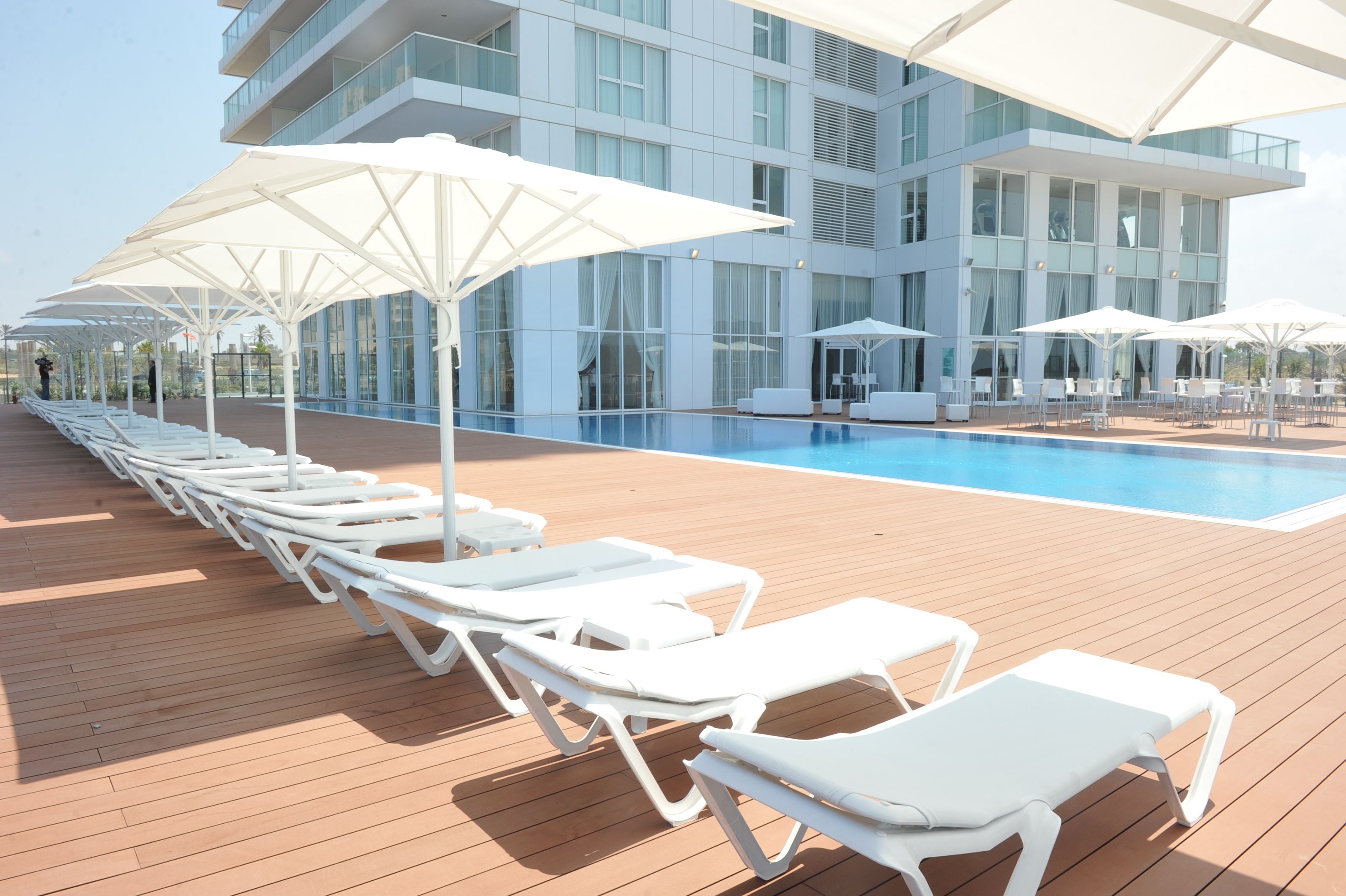 A poolside area with white lounge chairs and umbrellas, a modern building with large windows, and a clear blue pool.