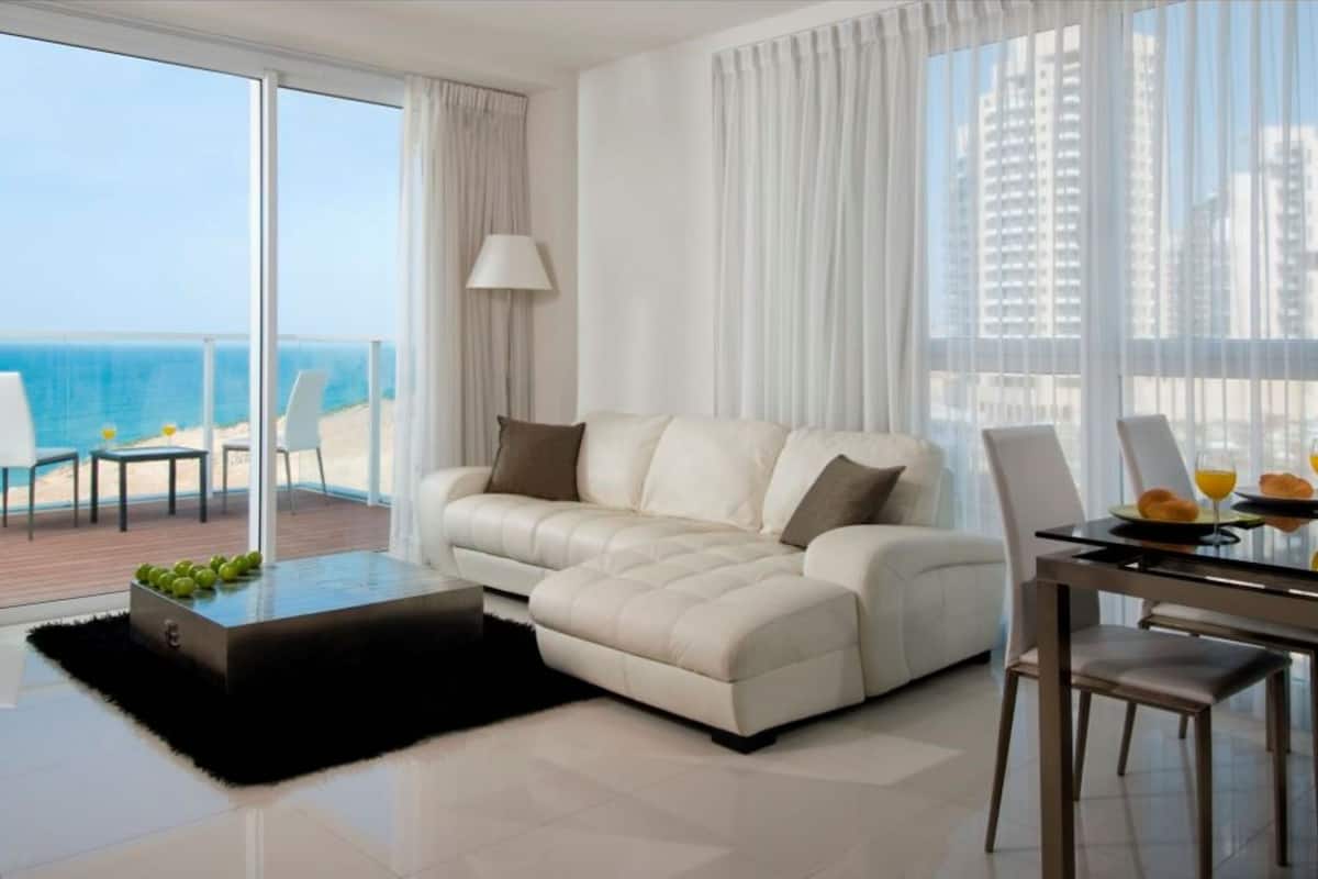 A modern living room with a sofa, a dining table with chairs, and a view of the ocean.