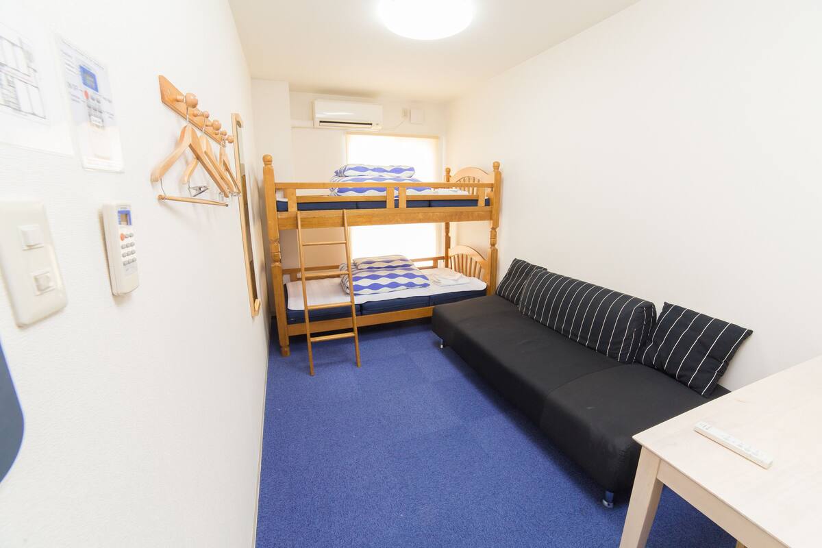 En køjeseng med ternet dyne, en sort sofa med stribede puder, et lille bord og en vægmonteret varmeovn.