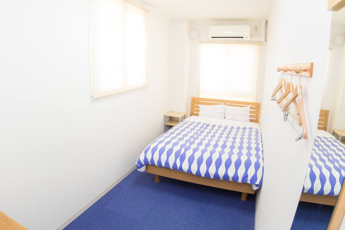 A bedroom with a single bed, a wooden headboard, a blue carpet, a window with blinds, and a wall-mounted air conditioner.