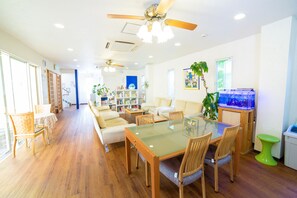 A spacious living area with a glass dining table, wooden chairs, a sofa, and a bookshelf.