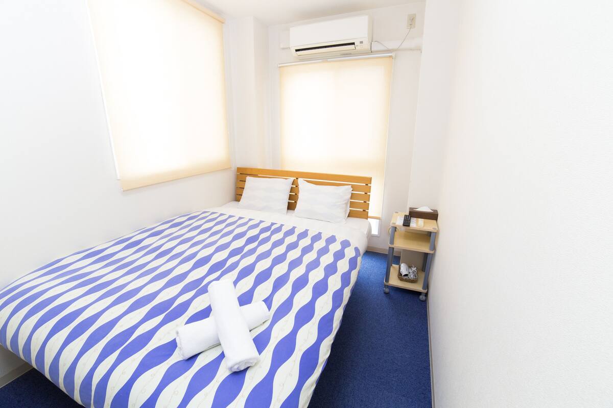 A hotel room with a bed, a nightstand, a shelf with toiletries, and an air conditioning unit.