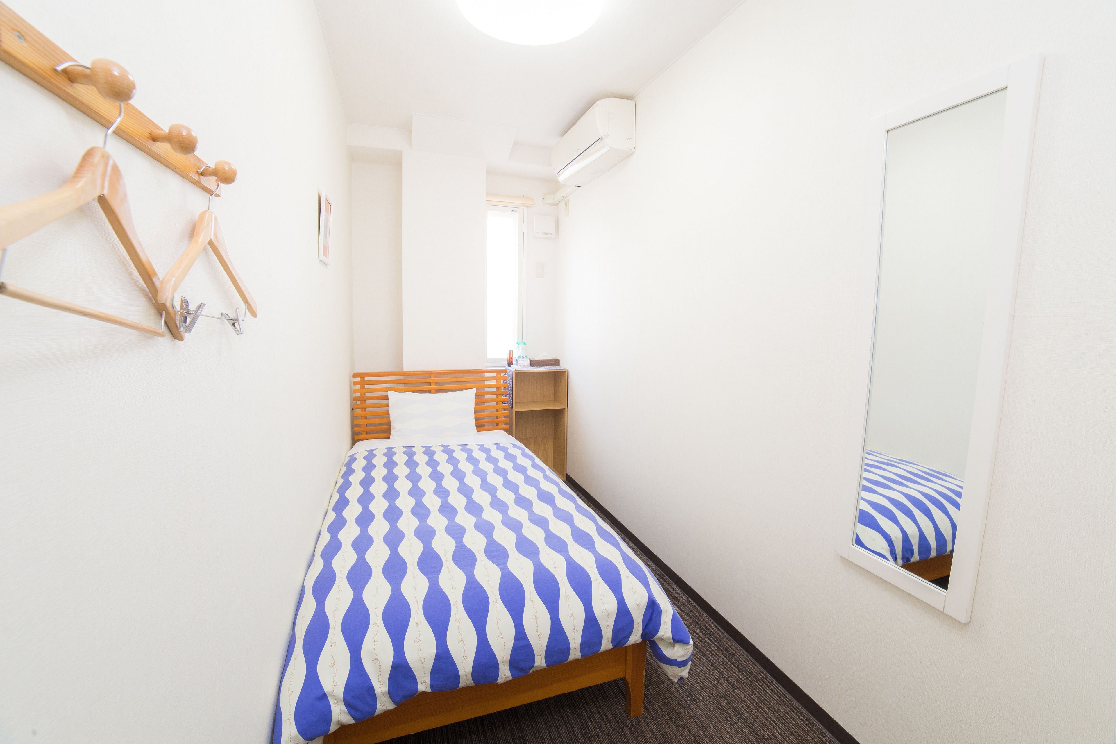A small, neatly arranged bedroom with a bed, a wooden headboard, a mirror, and a wall-mounted coat rack.