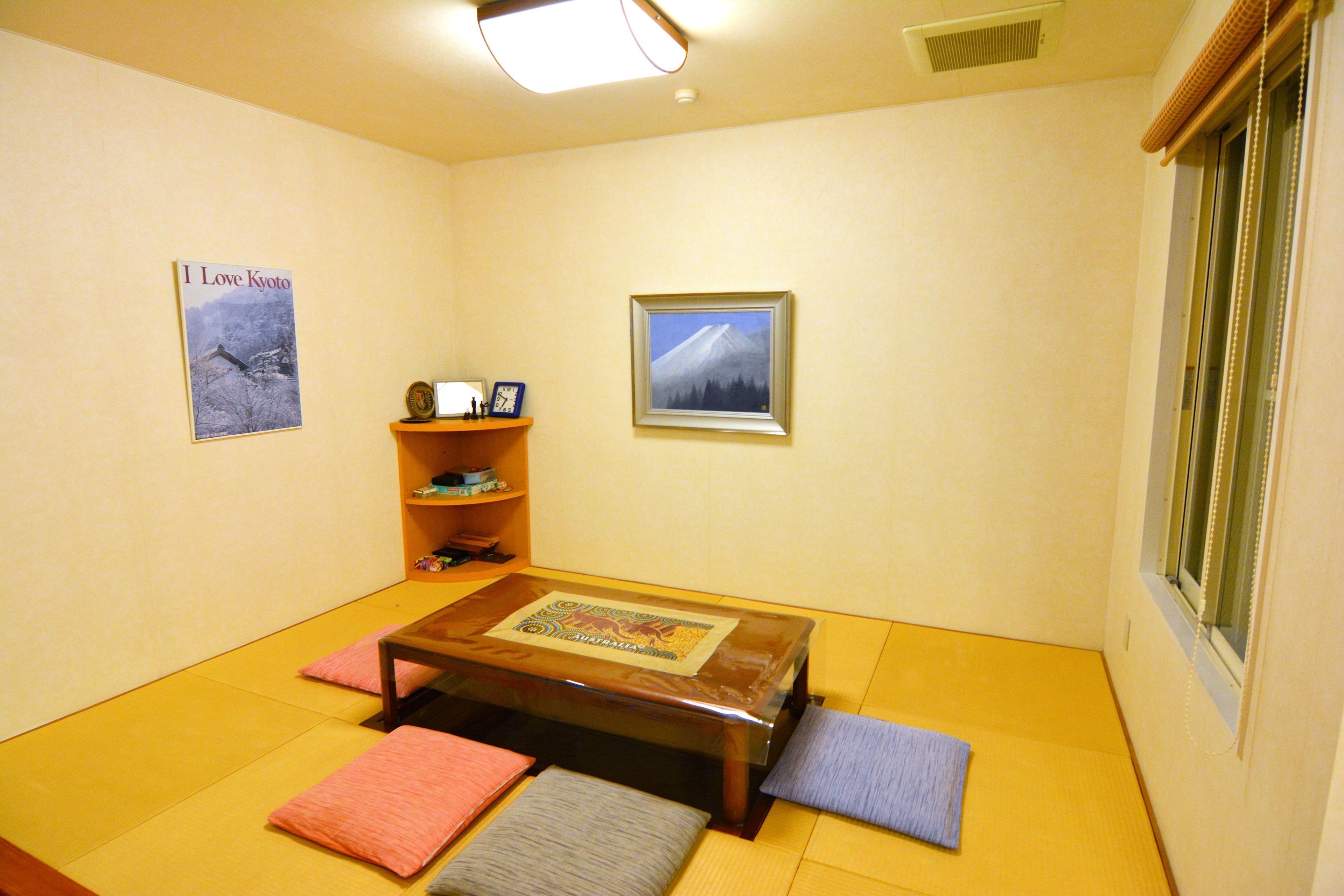 Ein Zimmer mit Tatami-Boden, einem niedrigen Holztisch mit Kissen, einem Bücherregal und zwei gerahmten Bildern an den Wänden.