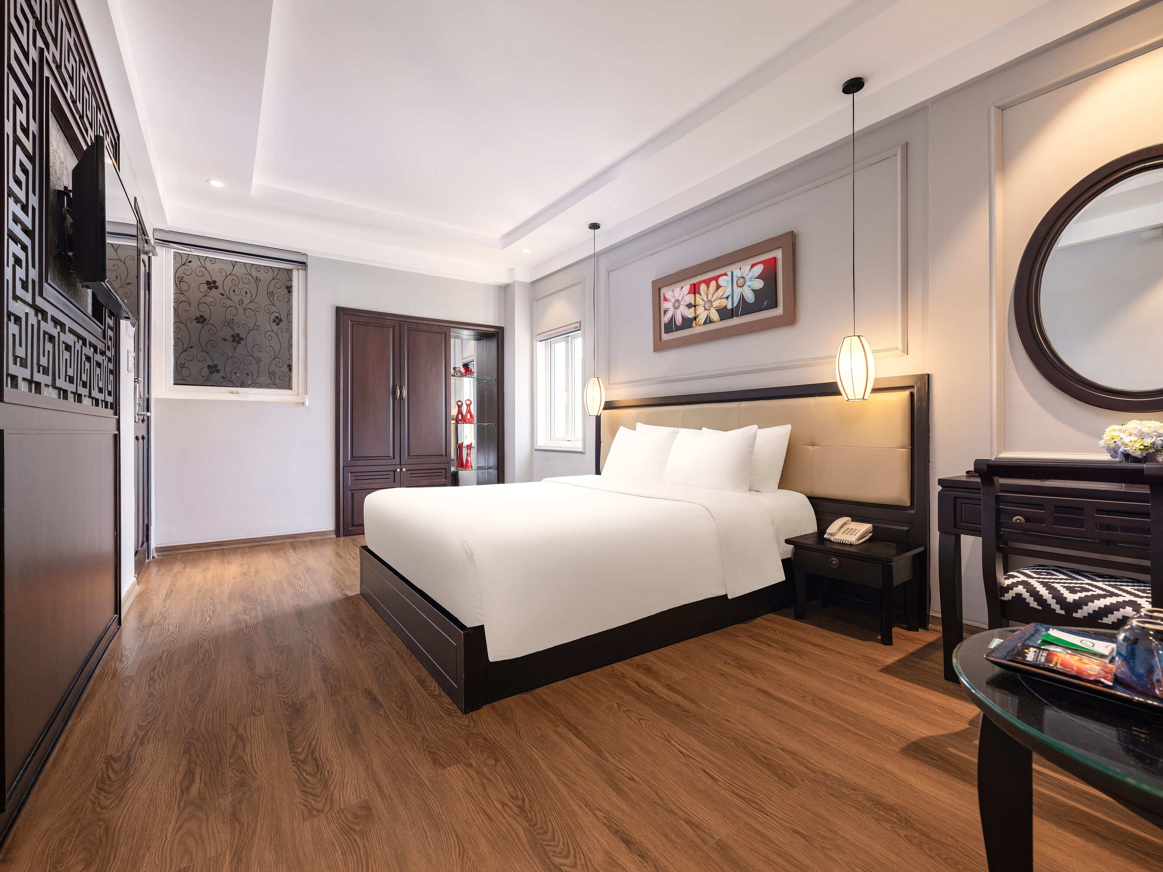A modern hotel room with a large bed, a bedside table with a lamp, a desk with a chair, and a decorative mirror.