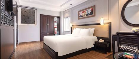 A modern hotel room with a large bed, a bedside table with a lamp, a desk with a chair, and a decorative mirror.