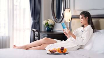 A person reading a book in a hotel room with a bed, a nightstand, a vase with flowers, and a lamp.