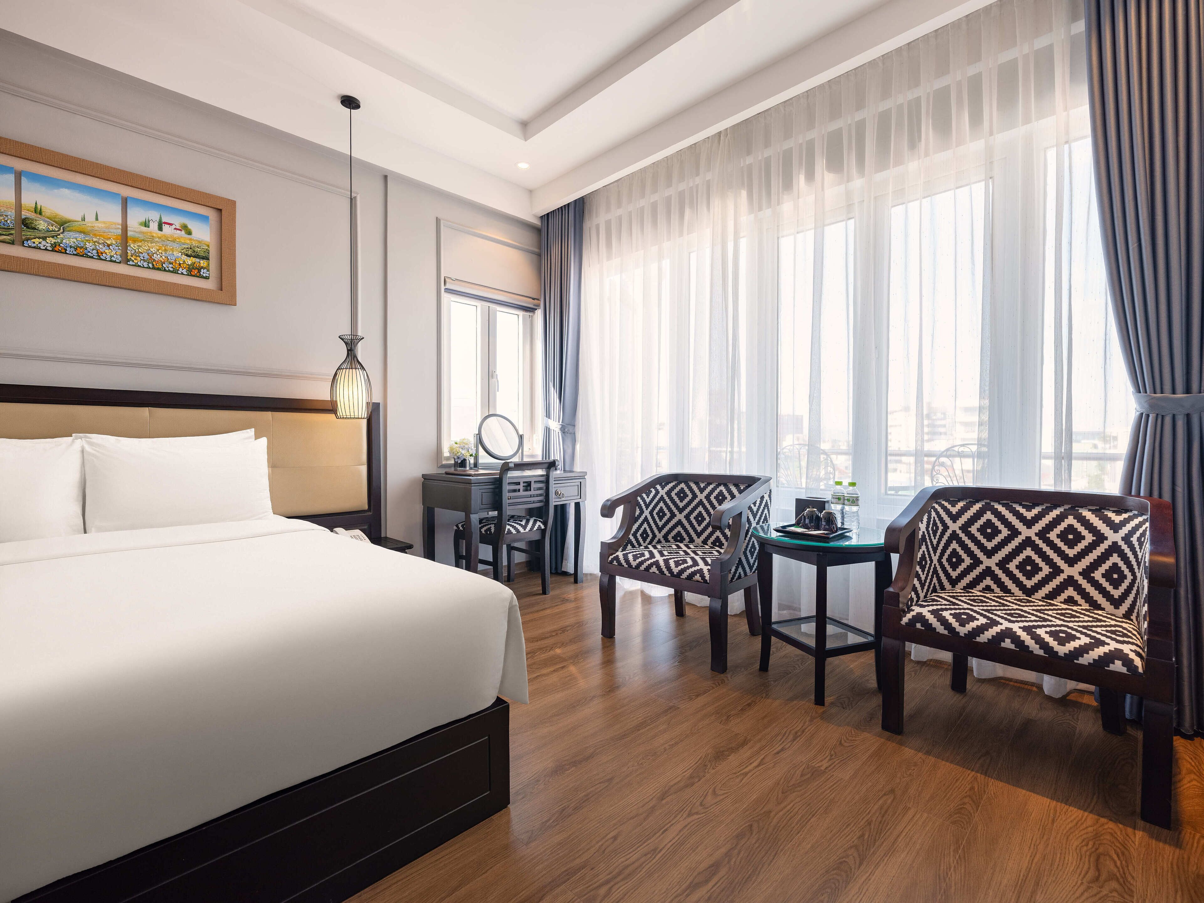 Une chambre d’hôtel avec un grand lit, un bureau avec une chaise, une petite table avec une chaise et une fenêtre avec des rideaux.