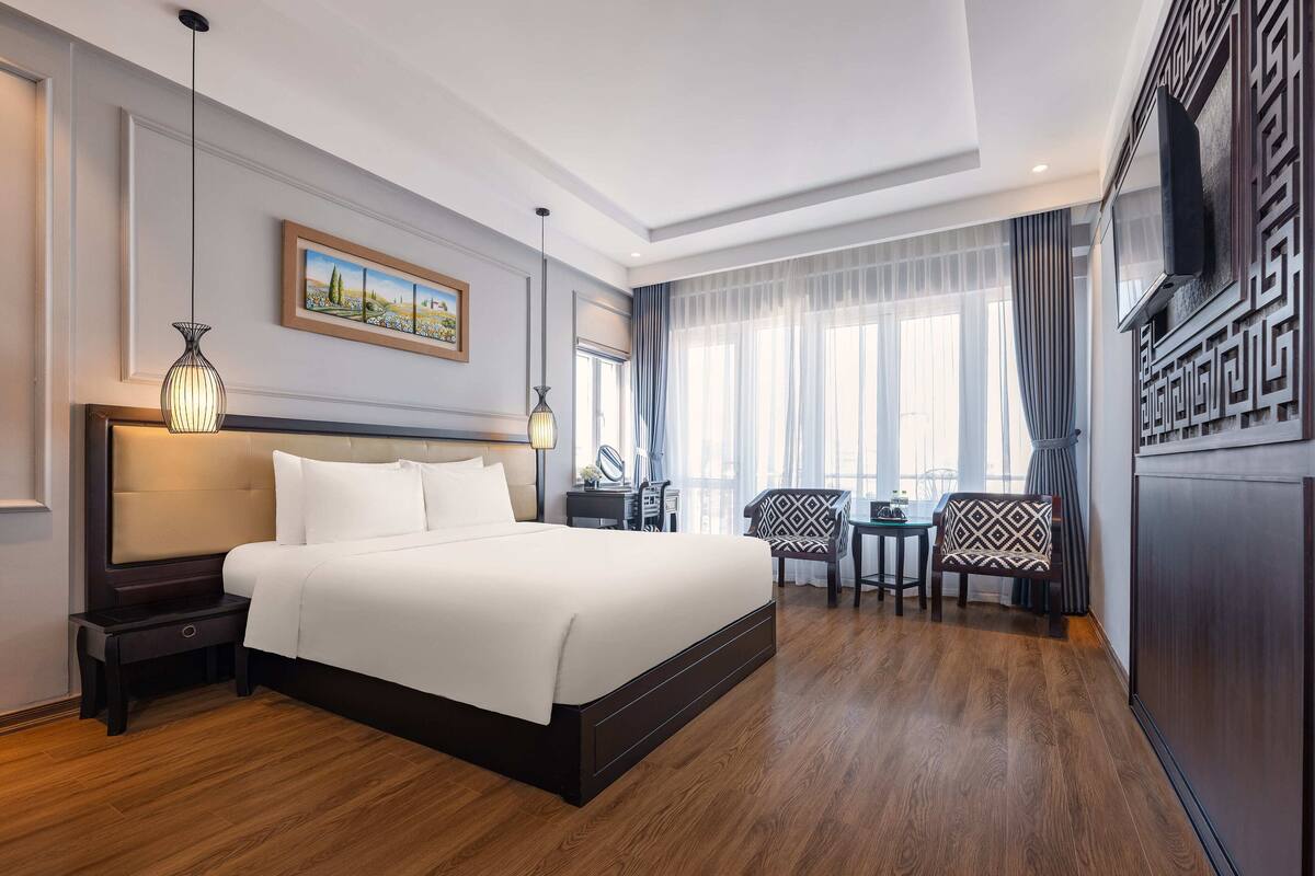 A hotel room with a large bed, bedside tables, a desk with chairs, a TV, and a window with curtains.