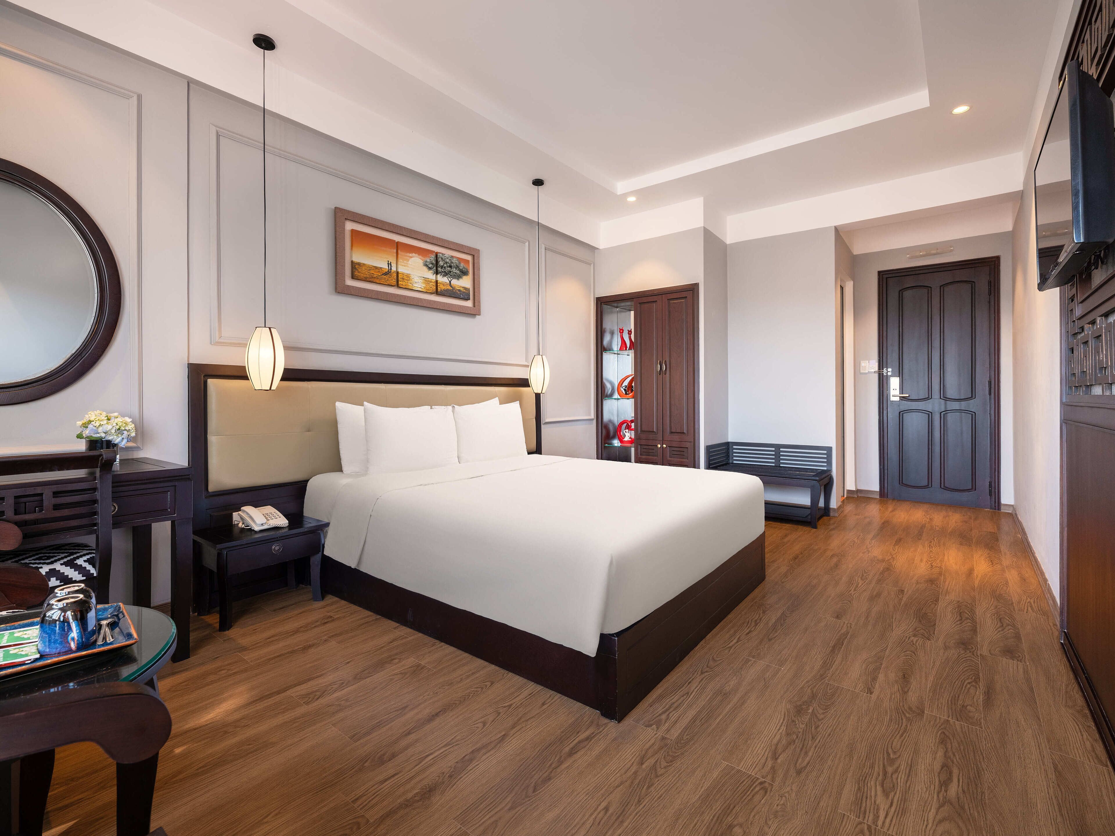 A hotel room with a large bed, a desk with a telephone, a chair, a bench, a wardrobe, and a painting on the wall.