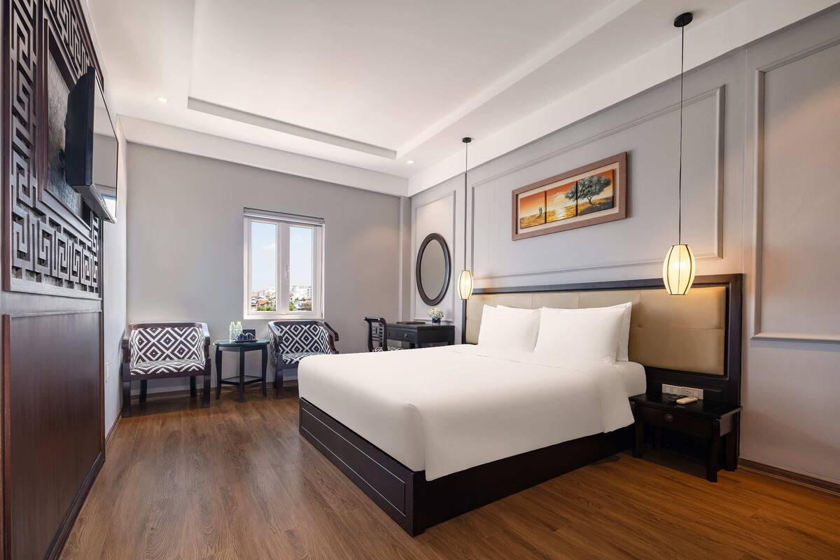 A hotel room with a large bed, a desk with a chair, a window with a view, and a painting on the wall.