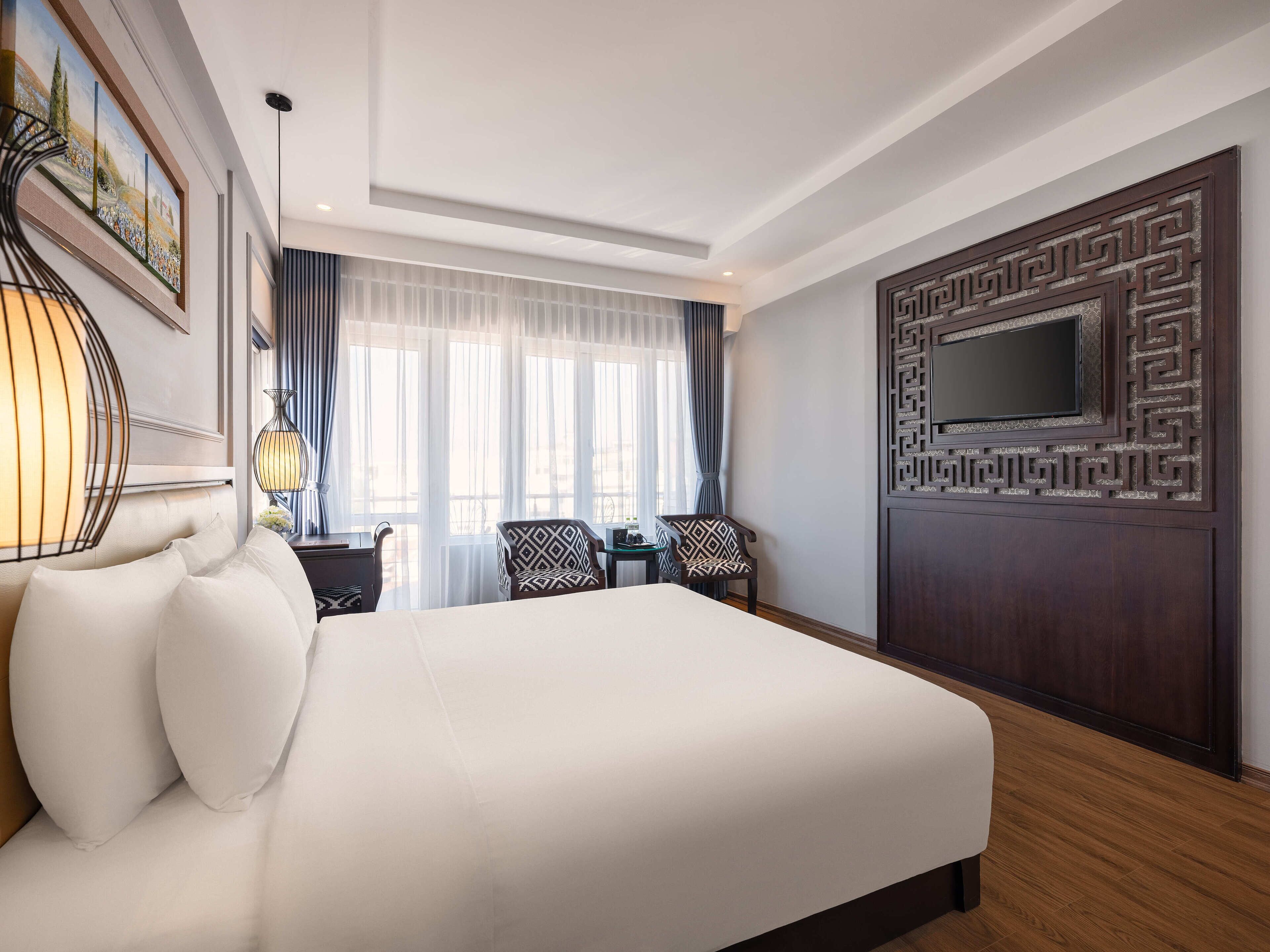 A hotel room with a large bed, two armchairs, a desk, and a TV mounted on a decorative panel.