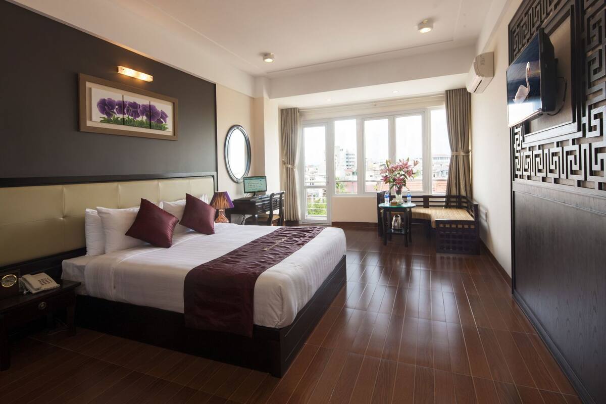 A hotel room with a large bed, a desk, a chair, a TV, and a balcony with a view.