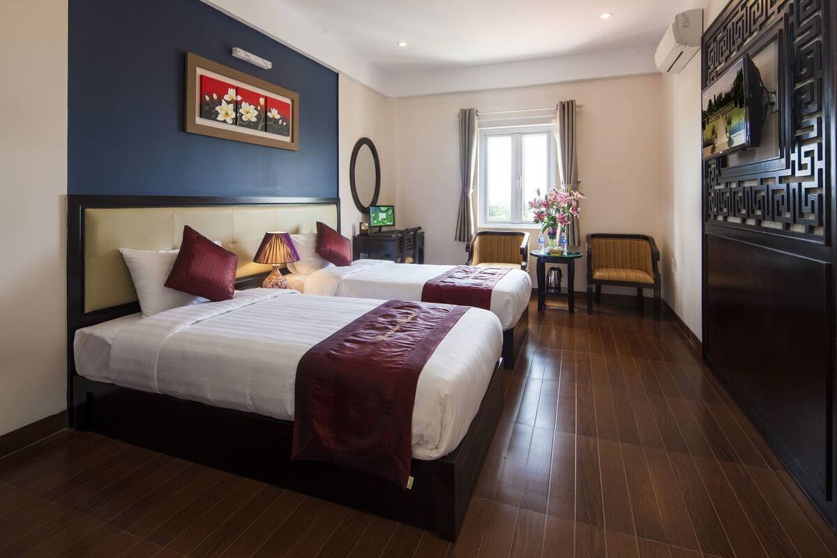 A hotel room with two beds, a TV, a small table with a vase, and a window with curtains.