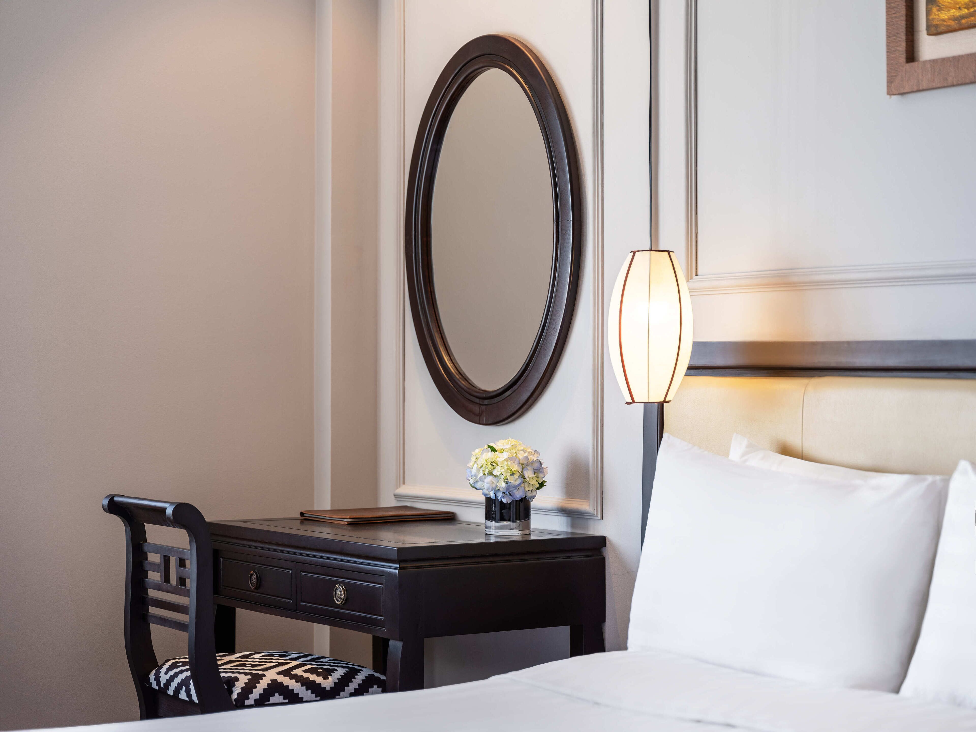 A hotel room with a bed, a desk with a chair, a mirror, a lamp, and a vase with flowers.