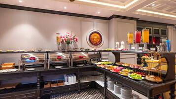 A buffet setup with various food items and beverages, a decorative wall with flowers, and a ceiling fan.