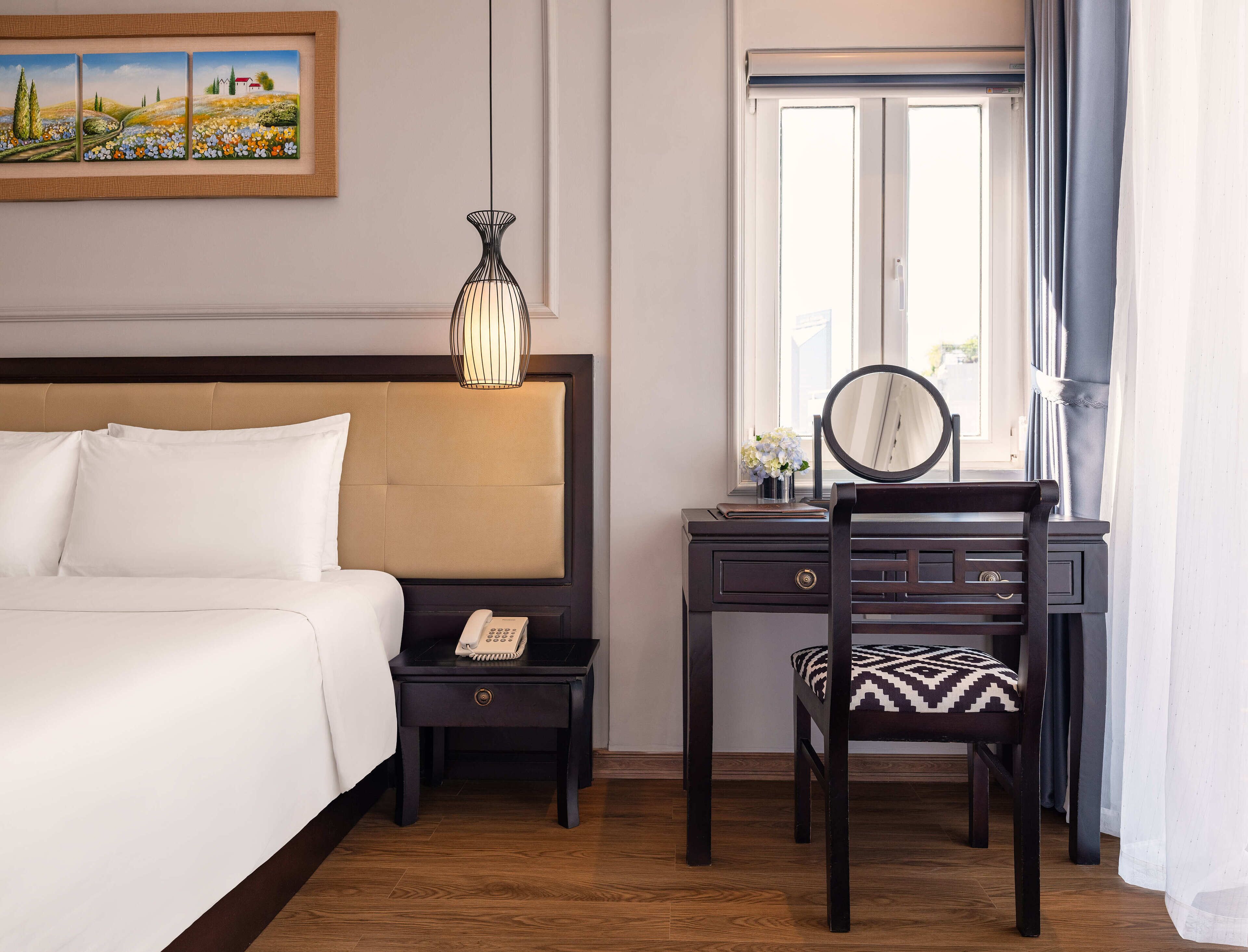 A hotel room with a bed, bedside table with a phone, a desk with a chair, a mirror, a window with curtains, and a painting on the wall.