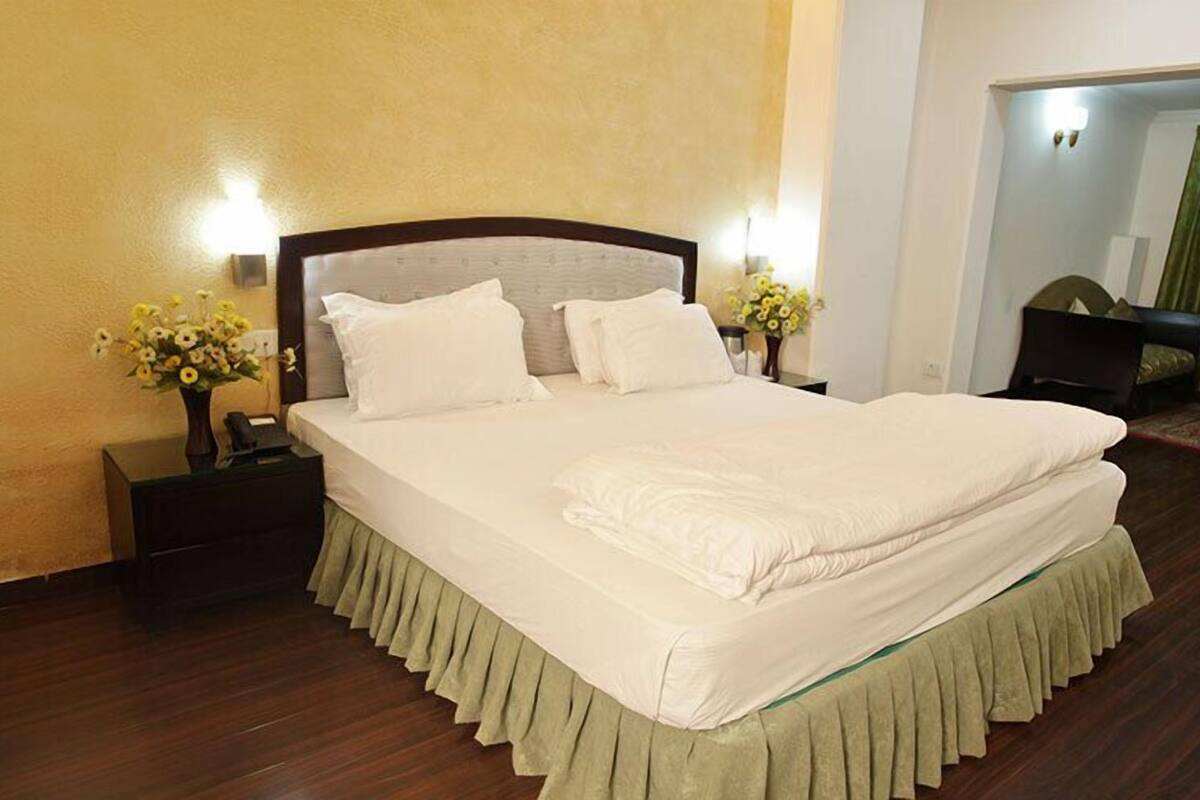 A neatly made bed with white linens, a wooden headboard, and a green skirt. There are two bedside tables with lamps and flower arrangements.