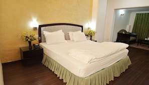 A neatly made bed with white linens, a wooden headboard, and a green skirt. There are two bedside tables with lamps and flower arrangements. - Hotel Grand Mumtaz Towers (Srinagar)