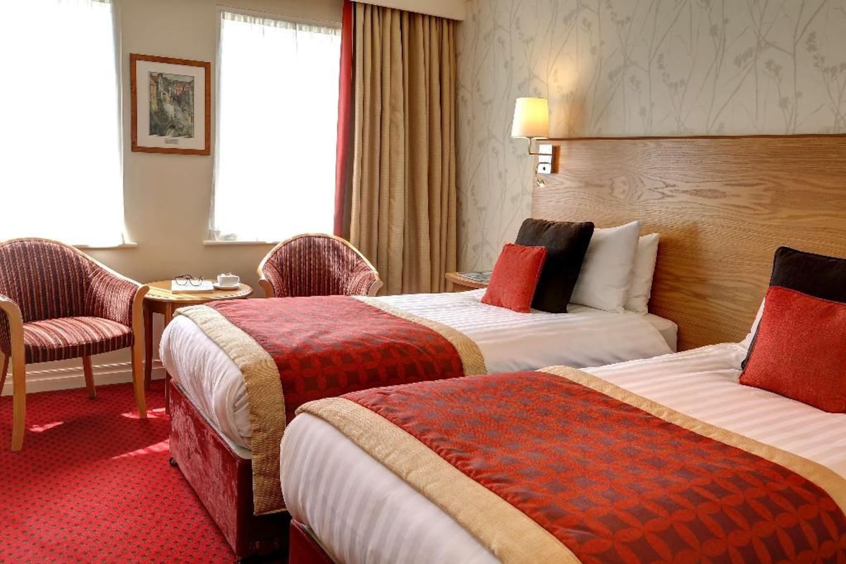 A hotel room with two beds, a sitting area with a chair, a small table, and a lamp.