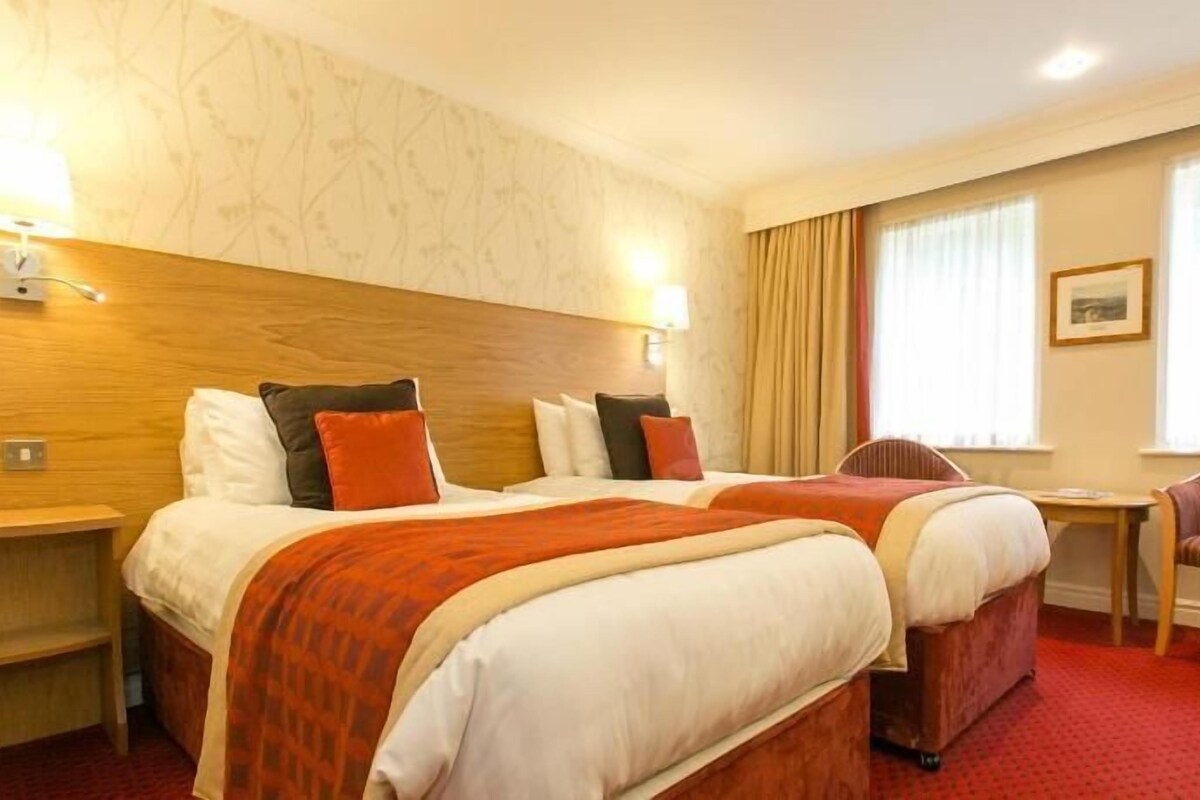 A hotel room with two beds, a small table, a chair, and a window with curtains.