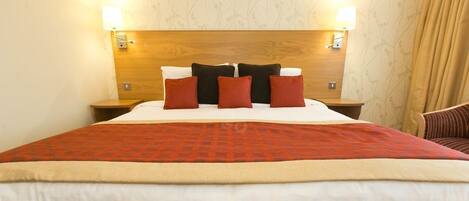 Habitación de hotel con cabecera de madera, una cama grande con colcha roja y beige, y dos lámparas de noche.
