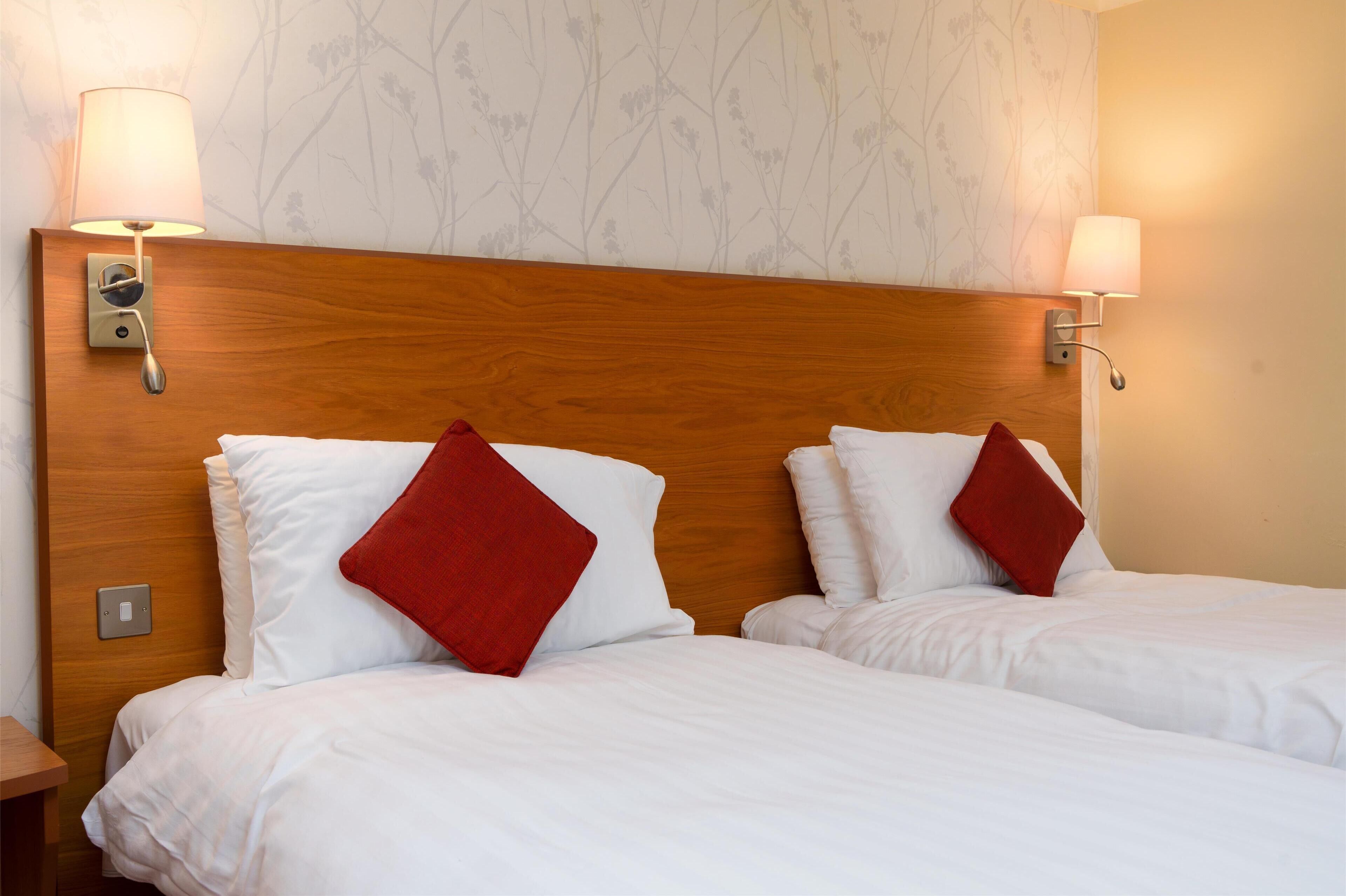 A hotel room with two beds, each with white bedding and red pillows, wooden headboard, and wall-mounted lamps.