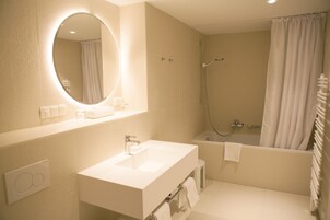 Uma casa de banho moderna com um espelho oval grande, uma sanita branca, um chuveiro e uma banheira.