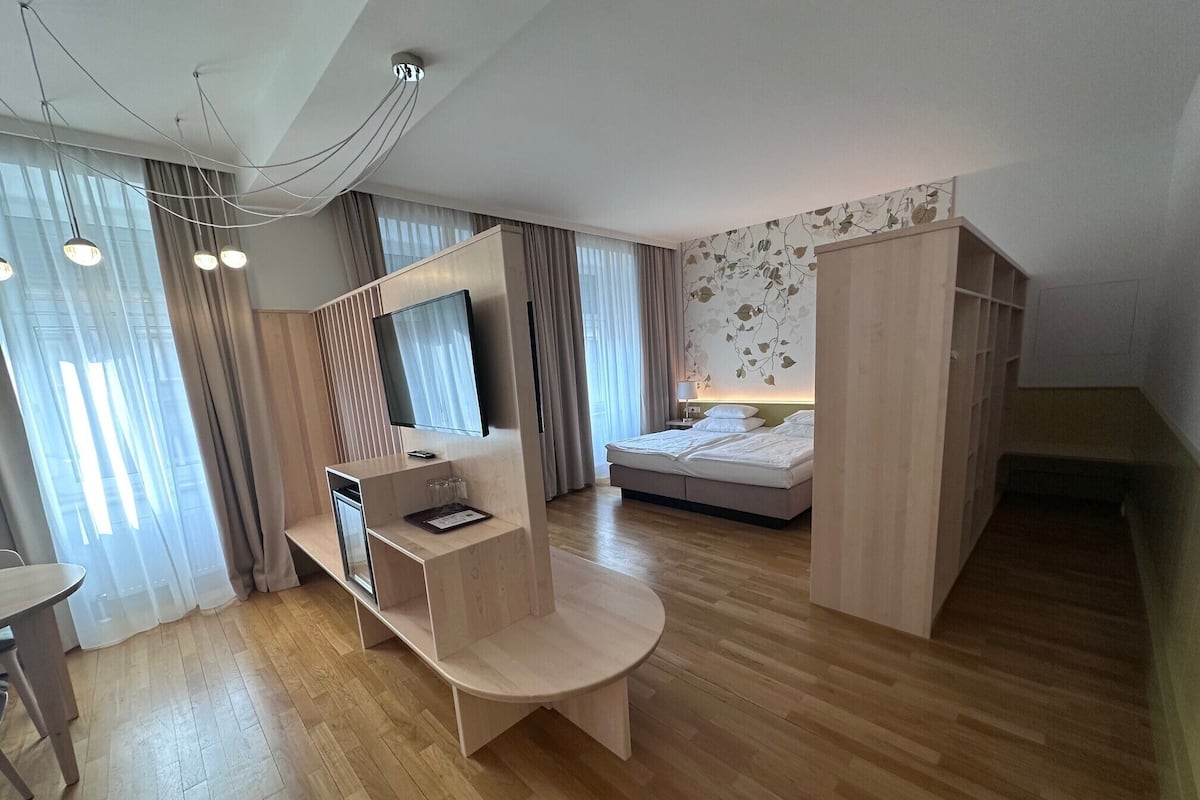 Una habitación de hotel moderna con suelo de madera, una cama, un escritorio con silla, un televisor y un armario.