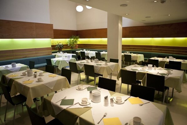 Un comedor con varias mesas redondas cubiertas con manteles blancos, sillas verdes y diferentes utensilios para comer.