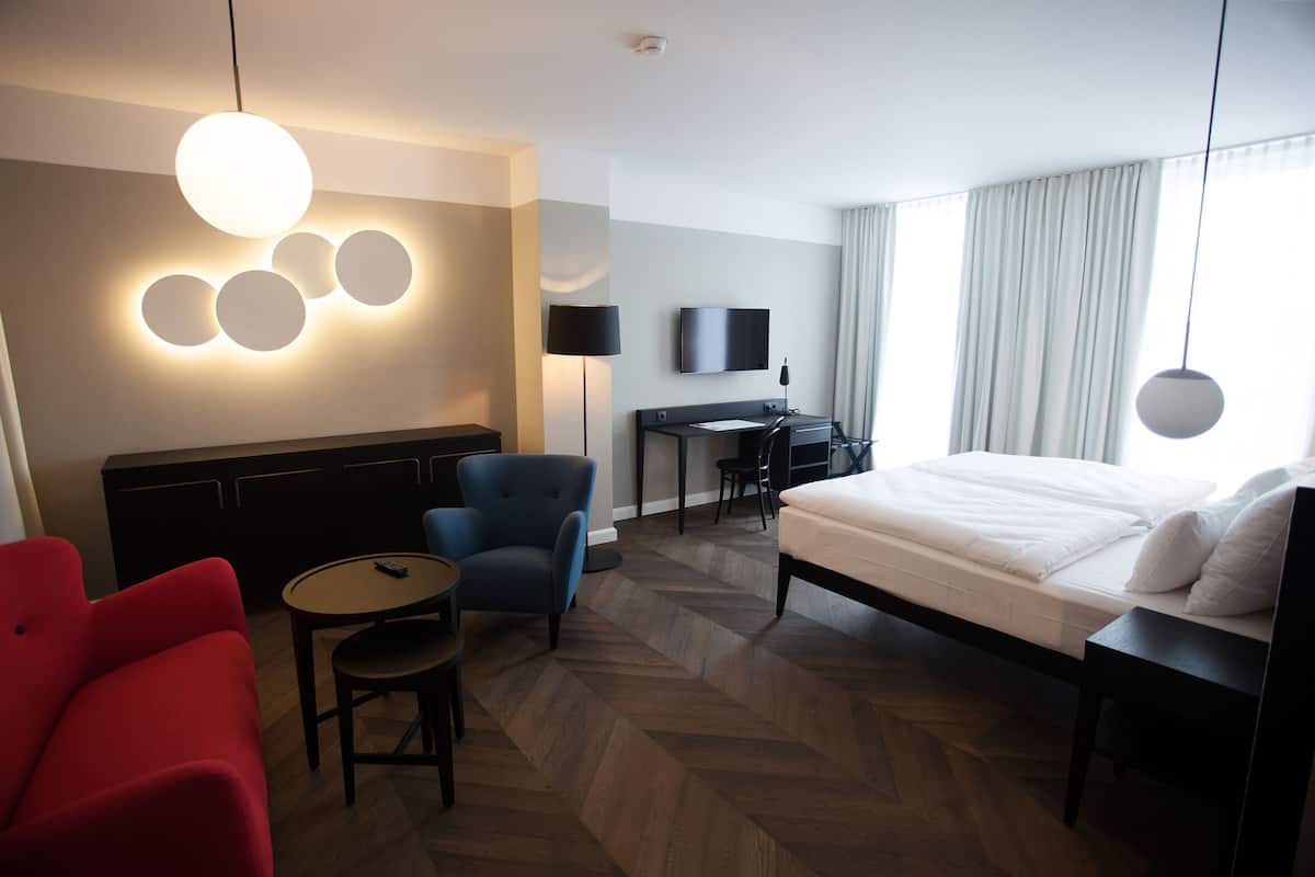Una habitación de hotel con una cama, un escritorio con silla, un televisor, un sillón rojo y una mesa redonda.