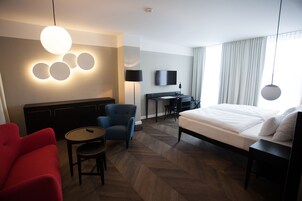 Una camera d'albergo con un letto, una scrivania con una sedia, una TV, una poltrona rossa e un tavolino rotondo.