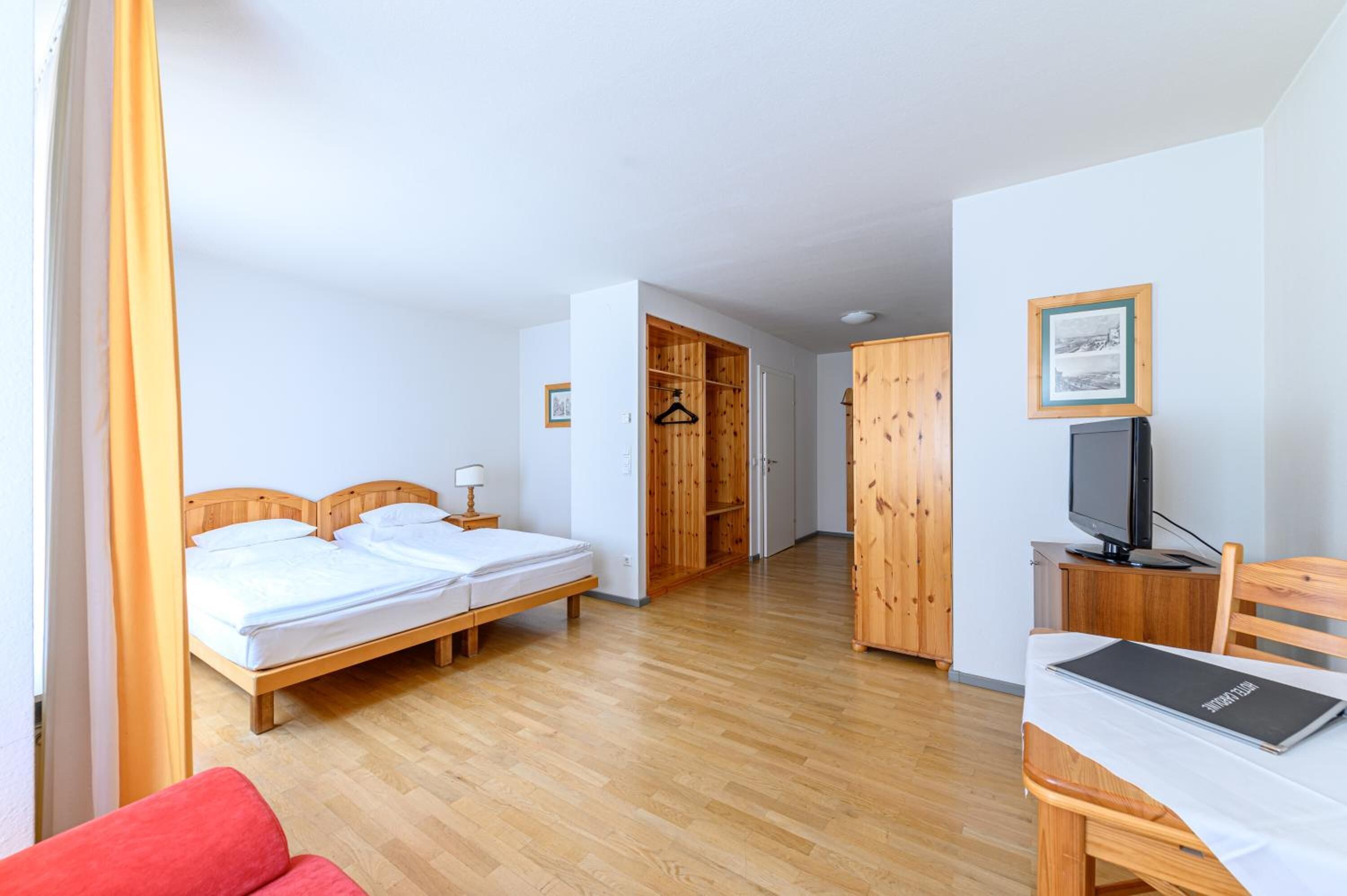 A hotel room with two beds, a wooden wardrobe, a television, and a desk with a laptop.