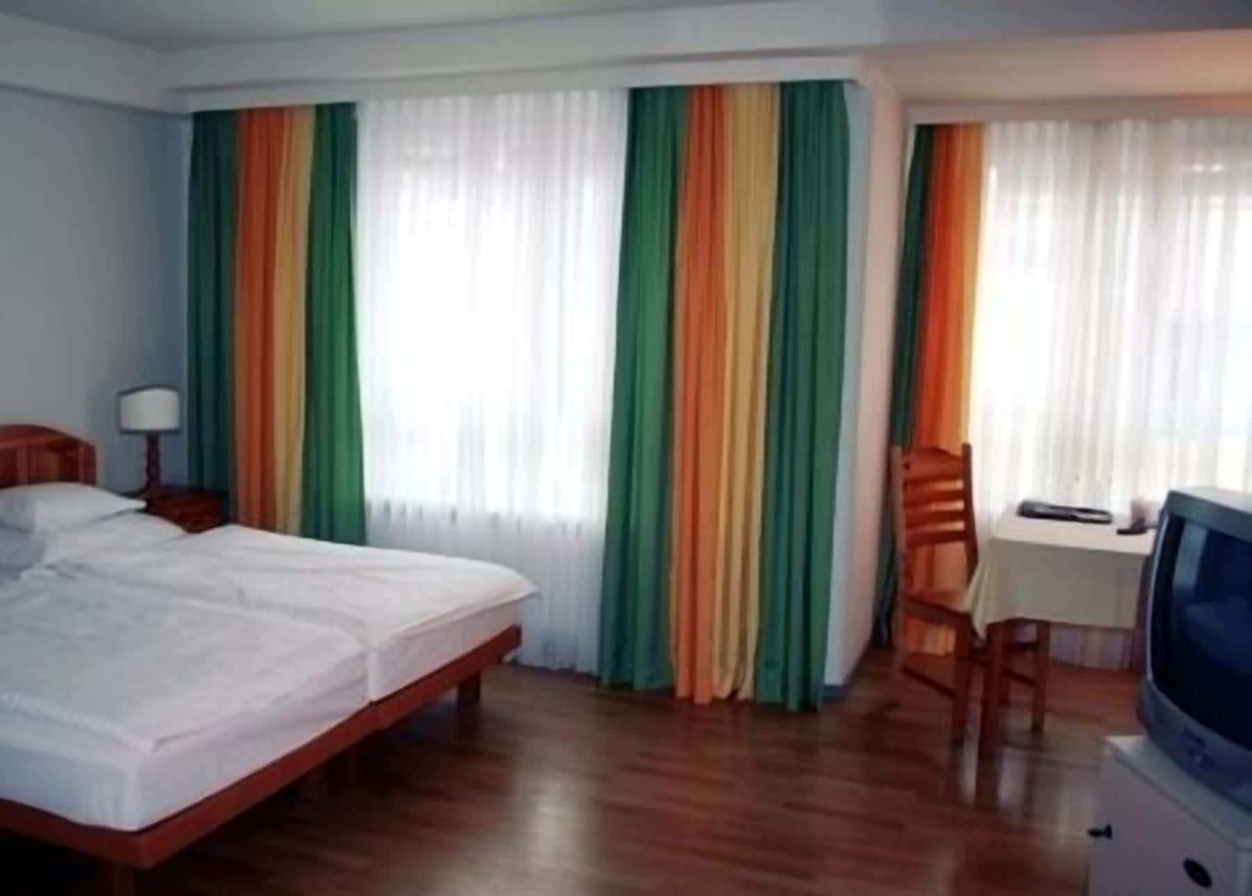 Una habitación de hotel con una cama, un escritorio con silla, un televisor y cortinas a rayas verticales.