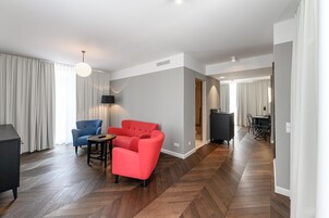 Quarto de hotel moderno com piso de madeira em padrão herringbone, um sofá vermelho, uma poltrona azul, uma pequena mesa redonda e um abajur preto.