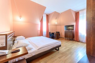 Quarto de hotel com cama, mesa de cabeceira, luminária, televisão e piso de madeira.