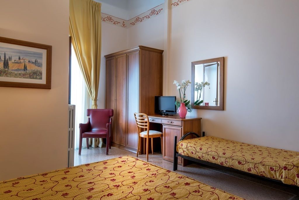 Ein Zimmer mit zwei Einzelbetten, einem Schreibtisch mit Computer, einem Stuhl, einem roten Sessel und einem Bild an der Wand.