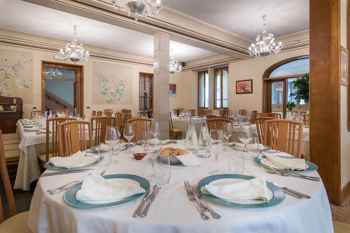 Una sala da pranzo formale con diversi tavoli apparecchiati per un pasto, con tovaglie bianche, piatti blu e posateria.
