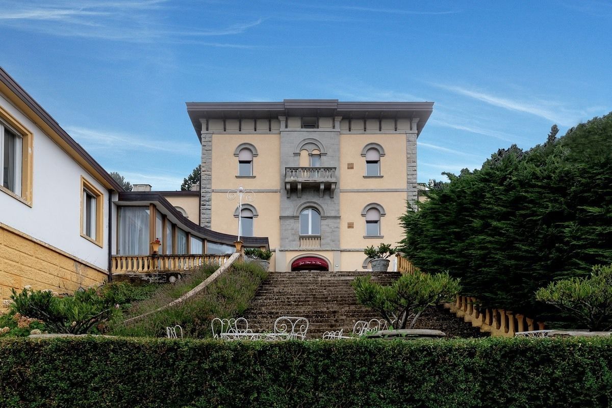 A multi-story building with a central entrance, a staircase leading up to it, and a well-maintained garden area with a hedge and metal railing.